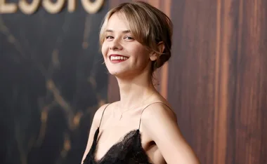 A person smiling in a sleeveless black lace dress, with a blurred dark background.