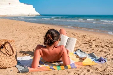 Woman sunbathes and reads a book on a towel at a sandy beach, with a woven bag nearby and the sea in the background.