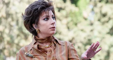 Person with short hair and patterned blouse making a gesturing hand motion outdoors.