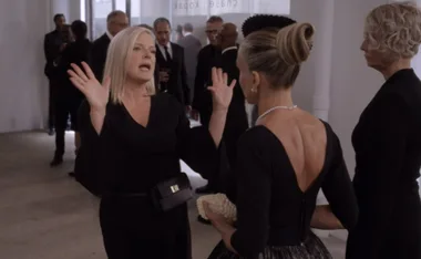 Two women in black dresses speaking animatedly at a formal event, surrounded by others in suits.