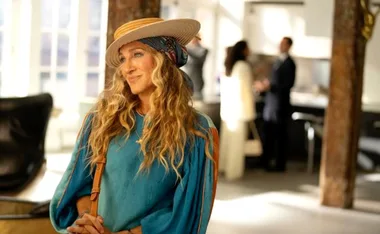 Woman in a stylish hat and blue blouse standing indoors, smiling softly. Two people chat in the blurred background.