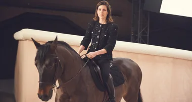 A woman in a black outfit rides a brown horse in an indoor arena setting.
