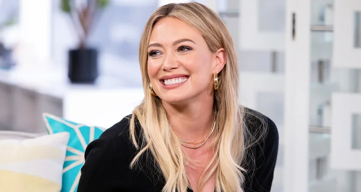 A person with long blond hair and gold earrings smiling indoors.