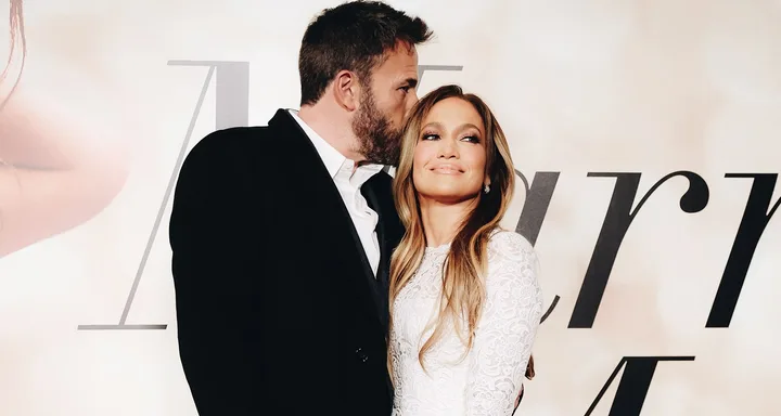 A couple embraces affectionately in front of a stylized backdrop with "Marry Me" text partially visible.