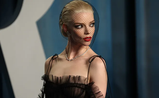 A woman with red lipstick and a netted veil wearing a black dress and necklace, looking to the side at a formal event.