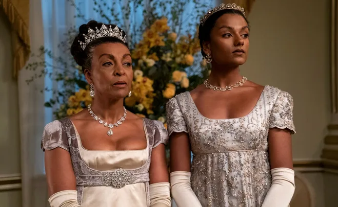 Two elegantly dressed women in period costume with jeweled tiaras, standing in front of yellow flowers.
