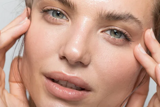Close-up of a person's face with clear skin, light makeup, and hands touching the temples.
