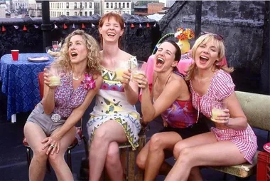 Women laughing and holding drinks on a rooftop, dressed in colorful summer outfits.