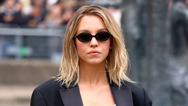 A woman with shoulder-length blond hair wearing black oval sunglasses and a black jacket.
