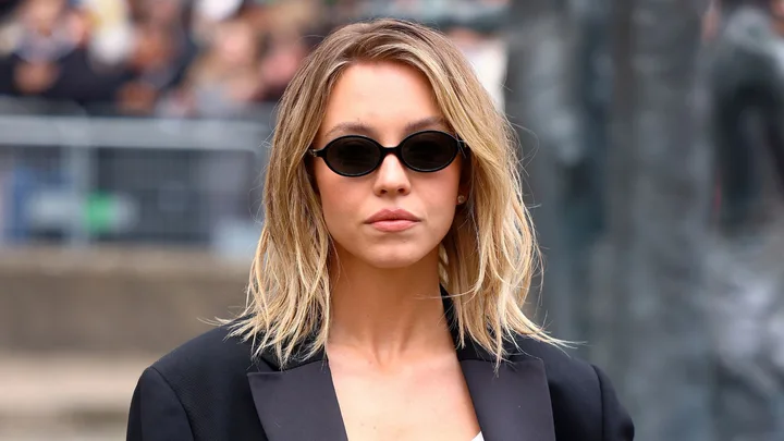 A woman with shoulder-length blond hair wearing black oval sunglasses and a black jacket.