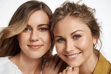 Two women smiling at the camera, one with wavy hair and the other with tied back hair, on a white background.