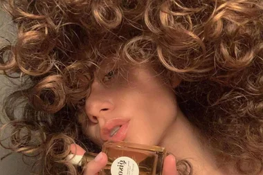 A person with curly hair holds a perfume bottle, partially obscuring their face.