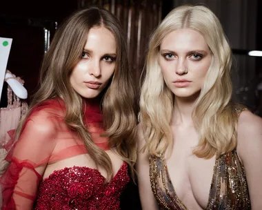 Models in elegant, detailed dresses preparing for a fashion show, one in red and the other in gold, indoors.