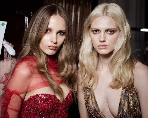 Models in elegant, detailed dresses preparing for a fashion show, one in red and the other in gold, indoors.