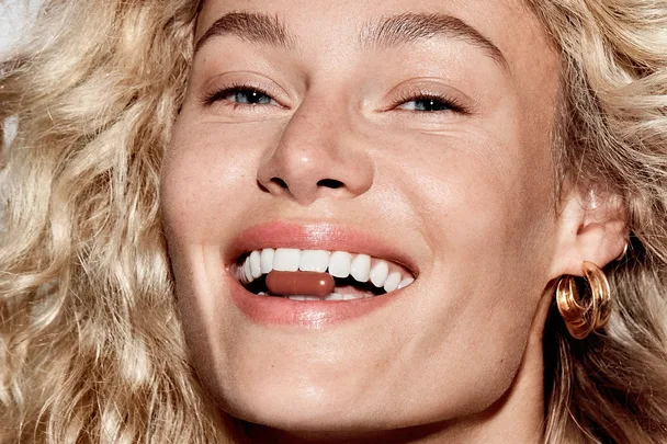 Smiling person with curly hair holding a capsule between teeth, wearing gold hoop earrings.