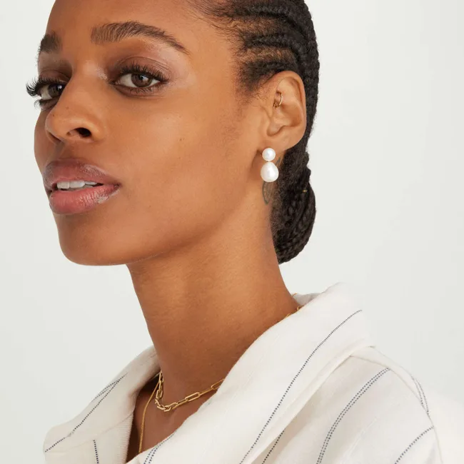 A woman with braided hair wears pearl earrings and a gold chain necklace, dressed in a white striped blouse.