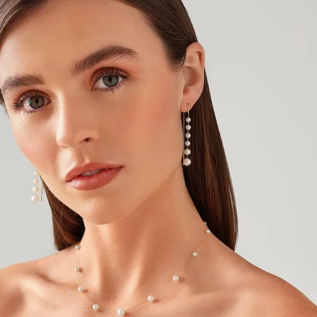 A woman with pearl earrings and necklace, minimal makeup, and straight brown hair poses against a plain background.