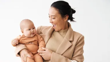 Woman in beige coat smiling, holding a baby in orange outfit against a white background.