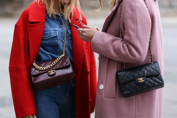 Two women in colorful coats, one red and one pink, each carrying Chanel handbags and using a smartphone.