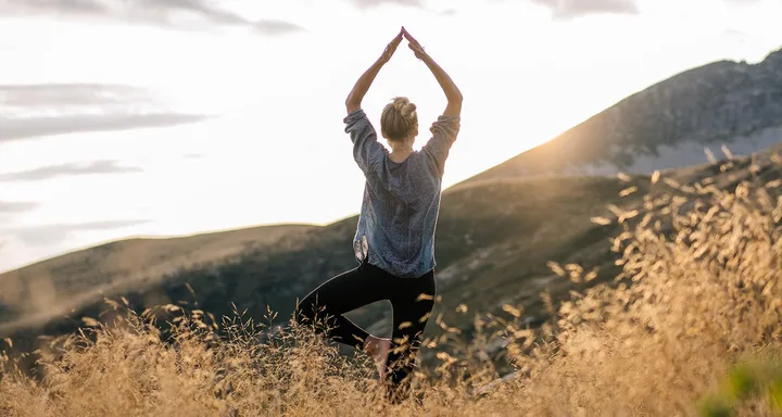 Person doing yoga in a grassy field with mountains in the background at sunset.