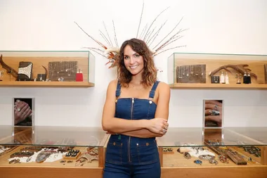 A woman in denim overalls smiles, arms crossed, standing in a jewelry store with displays of necklaces and rings.