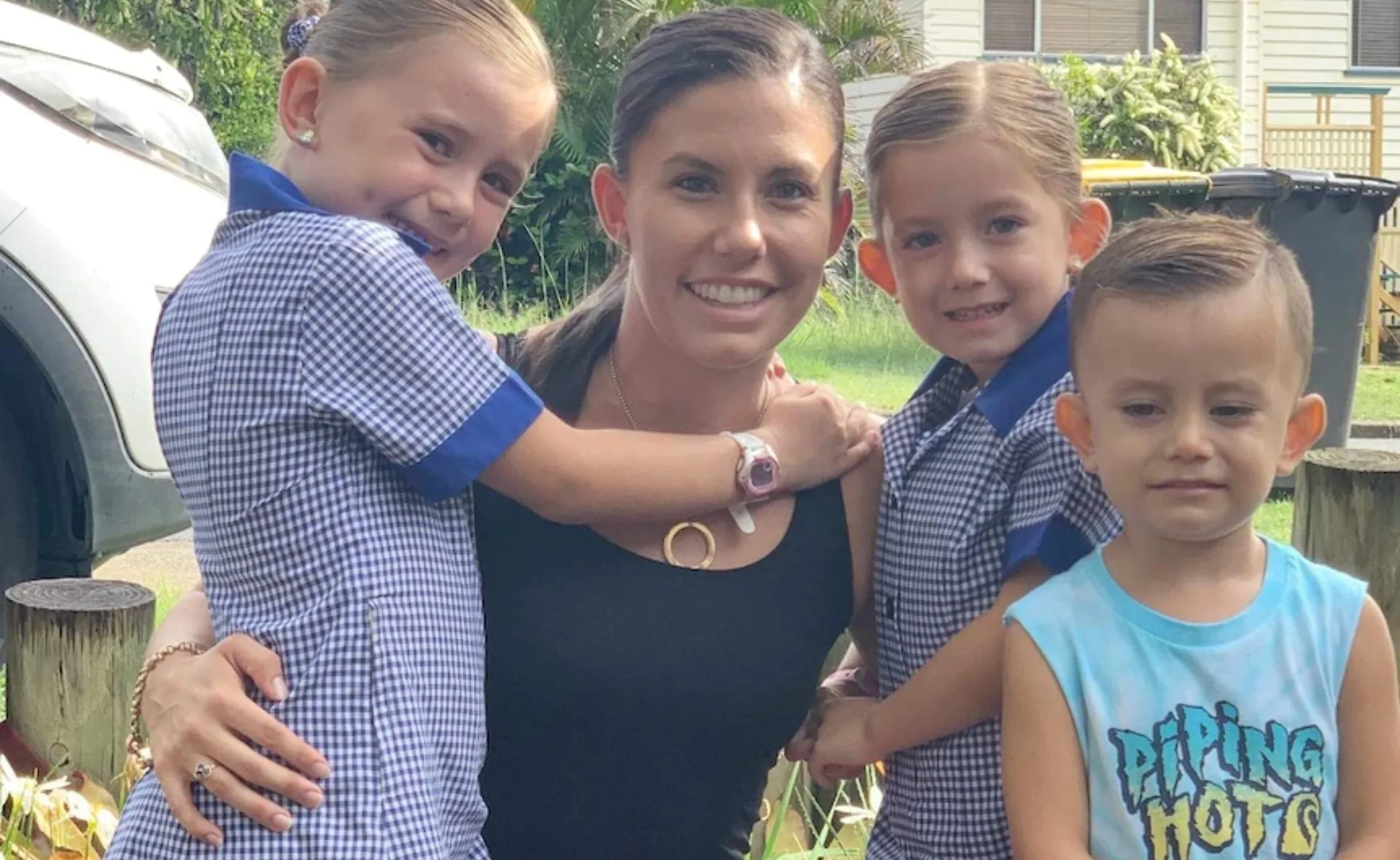 A woman smiles with three children outdoors, two in blue uniforms and one in a tank top.