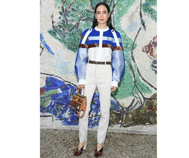 Woman in patterned top and striped pants poses in front of a mosaic wall, holding a patterned handbag.
