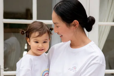 Mother and child in matching white shirts with rainbow designs, smiling in front of a window indoors.