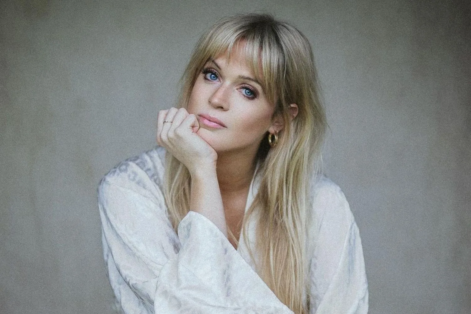 A woman with long blonde hair and bangs, wearing a light top, rests her chin on her hand, looking thoughtfully at the camera.