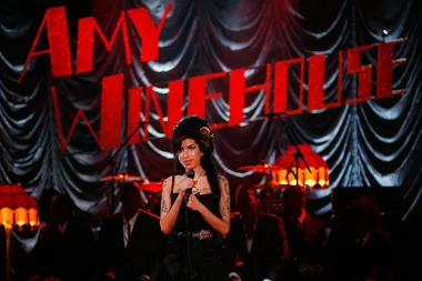 Singer performing on stage with "Amy Winehouse" in red letters and a band seated in the background.