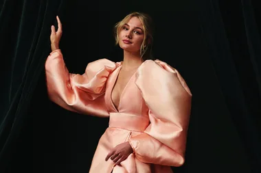 Alt text: Woman in a peach dress with puffy sleeves poses gracefully against a dark backdrop.