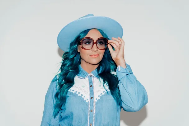 Person with turquoise hair, wearing a blue hat and denim shirt, adjusts oversized sunglasses against a light background.