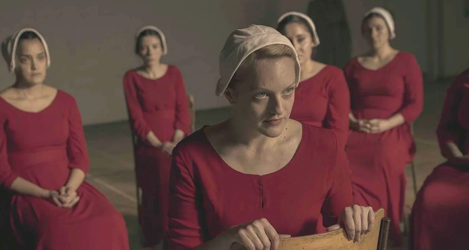Women in red dresses and white bonnets sitting; scene from "The Handmaid's Tale."