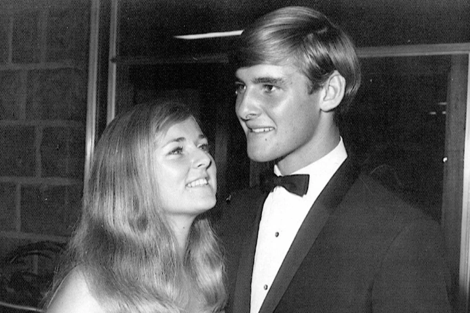 A young couple dressed formally, the woman looking up at the man who is wearing a tuxedo, both smiling.