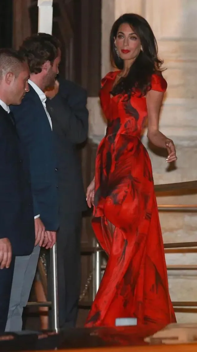 A woman in a red dress with a black pattern stands on steps, with two men in suits nearby.