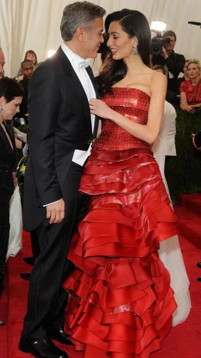 A couple dressed in formal attire smiles at each other on a red carpet, surrounded by photographers.