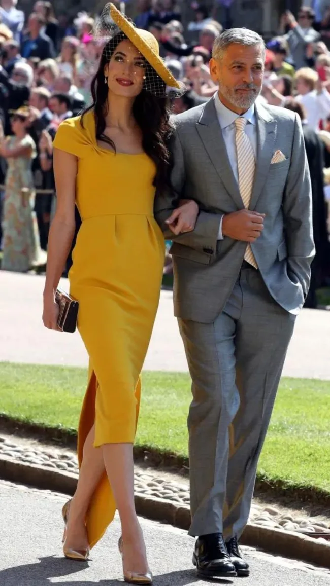 A man in a gray suit and a woman in a yellow dress and hat, walking arm-in-arm outdoors at an event.