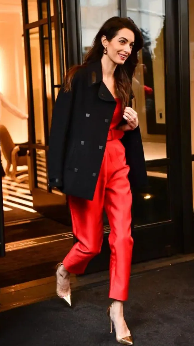 Woman in red outfit and black coat, smiling, walking outside a building, wearing gold heels.
