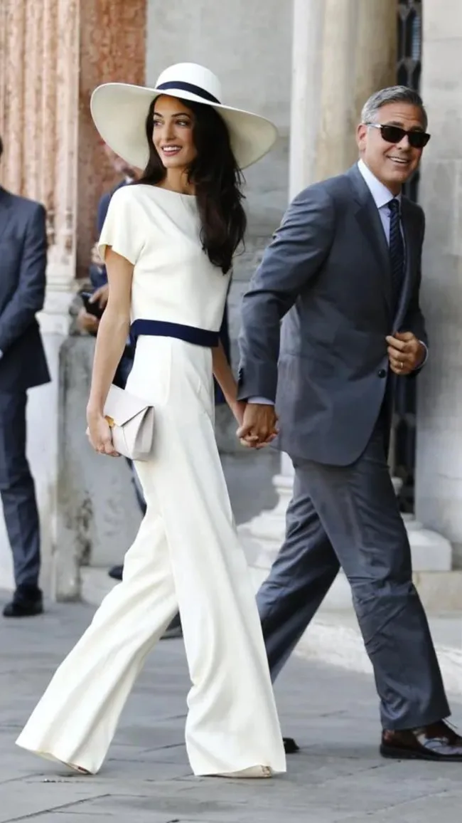 A couple walks hand-in-hand, dressed elegantly; the woman in a white outfit and hat, the man in a gray suit and sunglasses.