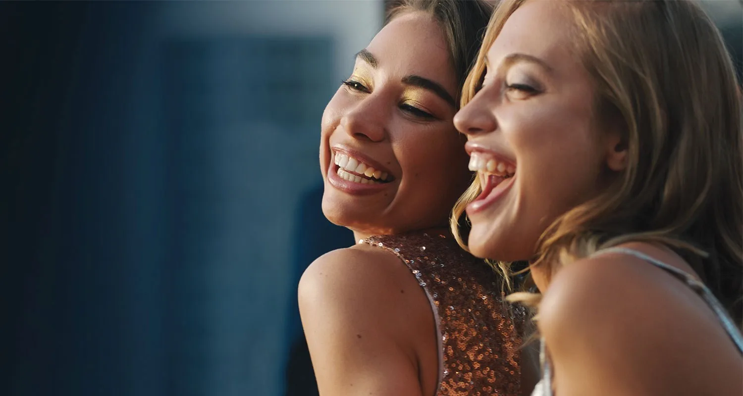 Two women smiling joyfully, wearing sequined tops, captured in a close-up photograph against a blurred background.