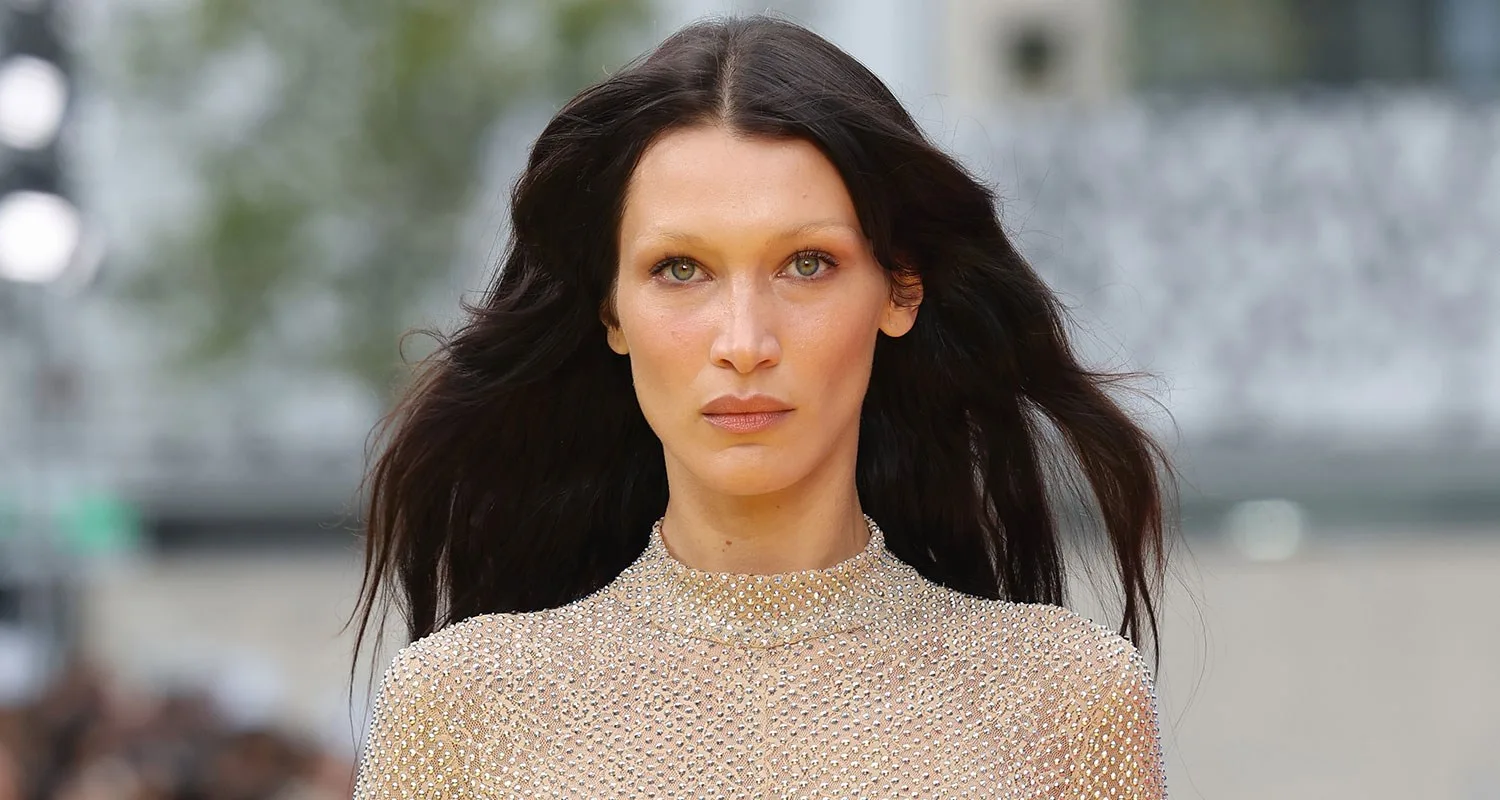 A model with long dark hair wears a sheer, shimmering top, walking outdoors.
