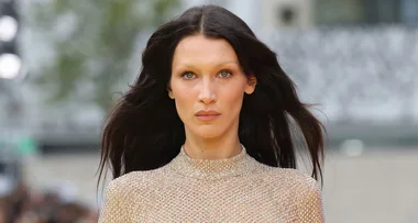 A model with long dark hair wears a sheer, shimmering top, walking outdoors.