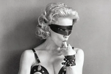 "Person in a black mask and studded outfit, licking a finger, looking sideways."