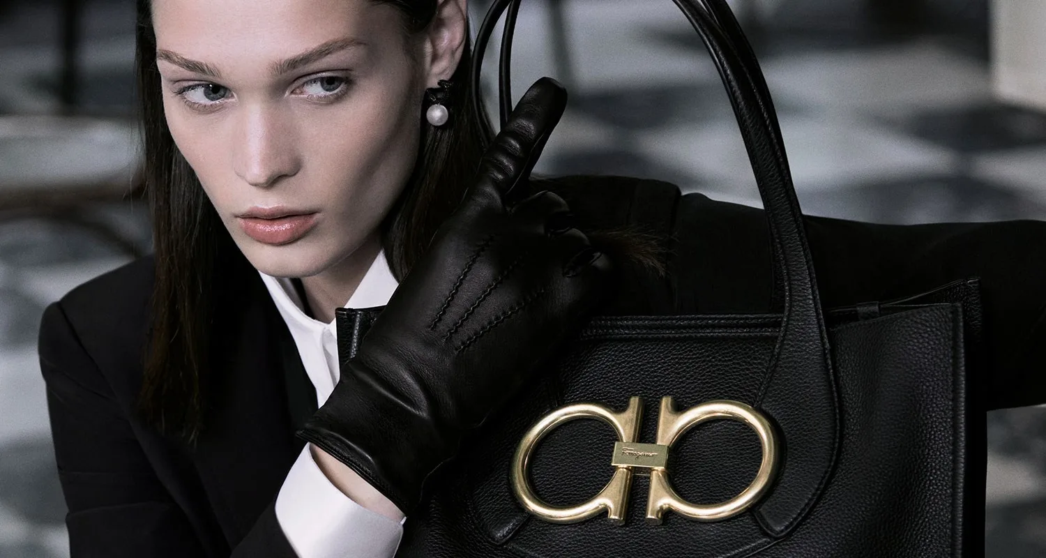 Woman in black suit with gloves holding a designer handbag with large gold logo.