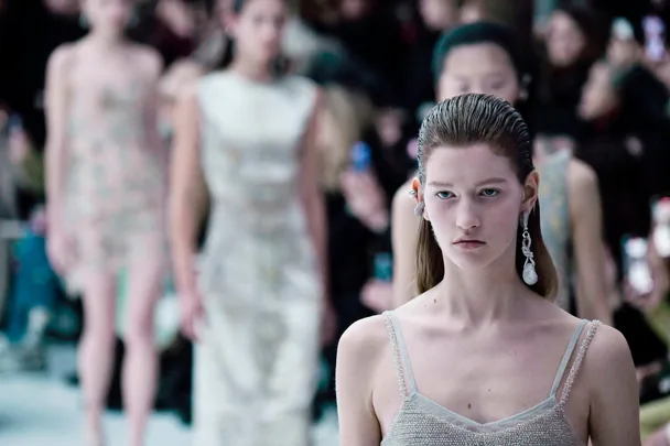 Models walking runway in elegant dresses at a fashion show, focused on front model with slicked-back hair and statement earrings.