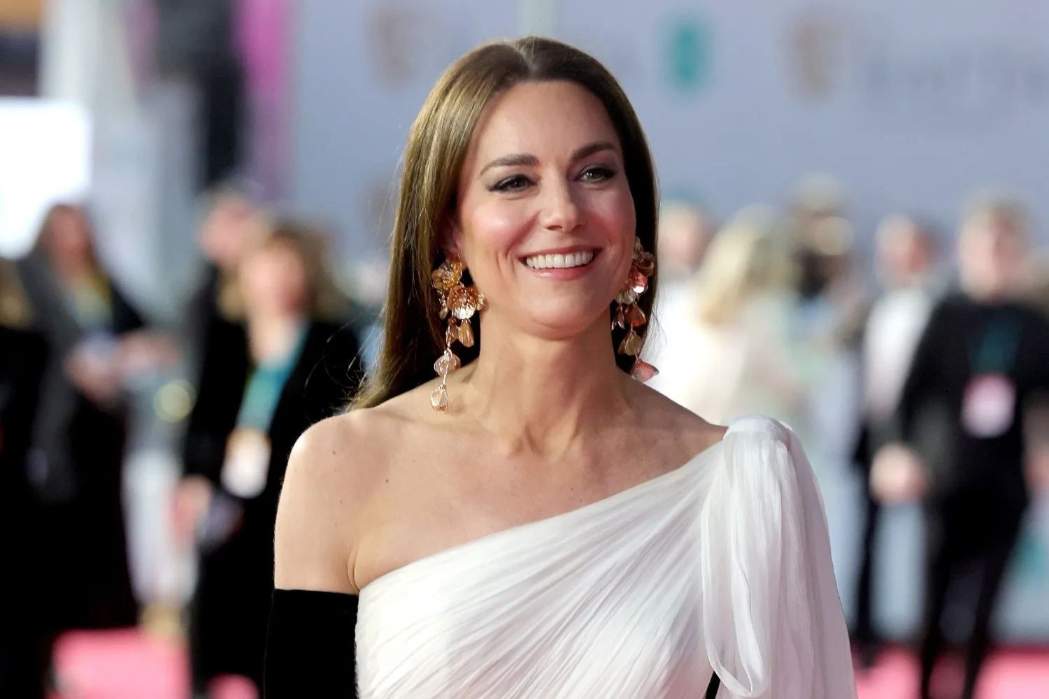 Woman in elegant white one-shoulder gown with long earrings, smiling at a formal event. Blurred crowd in background.