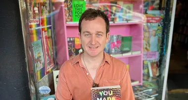 Man in orange shirt holding a book titled "You Made a Fool of Death with Your Beauty" in front of a bookstore window.