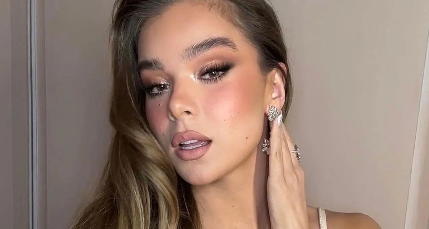 A woman with long hair, makeup, and jewelry poses while touching her ear, showcasing elegant earrings.