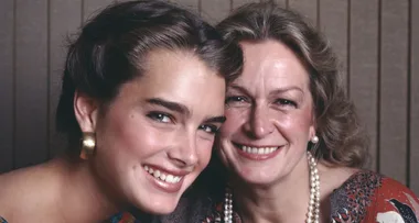 Two women smiling closely together, one younger with dark hair and the other older with light hair, both wearing earrings.