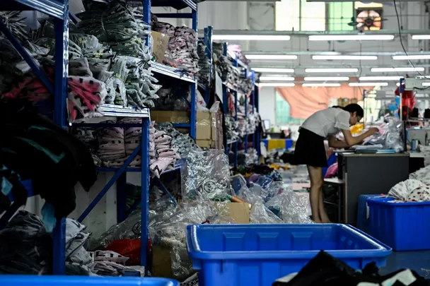 A person works in a garment warehouse, surrounded by shelves of folded clothes and plastic bins.